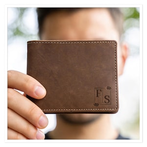 Könnte beinhalten: Braunes Lederportemonnaie mit weißen Nähten und einem Monogramm. Das Portemonnaie zeigt die Initialen "F S" in einem quadratischen Rahmen, akzentuiert mit Blatt- und Pfeildesigns. Das Portemonnaie wird vor einer Person gehalten.