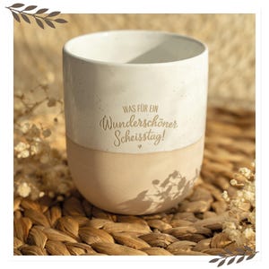 Könnte beinhalten: Zweifarbige Keramiktasse mit cremefarbenem Oberteil und beigefarbenem Unterteil. Die Tasse trägt den deutschen Text "WAS FÜR EIN Wunderschöner Scheisstag!" in einer Schriftart. Die Tasse steht auf einer gewebten Oberfläche.