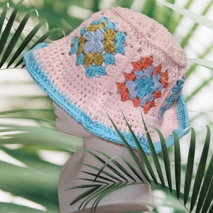 Può includere: Un cappello da sole rosa all'uncinetto con quadrati di nonna blu e arancioni. Il cappello ha un bordo blu.