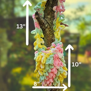 May include: A crocheted bag with a pink, green, and yellow color scheme. The bag is 13 inches tall and 10 inches wide. It is hanging from a tree branch.
