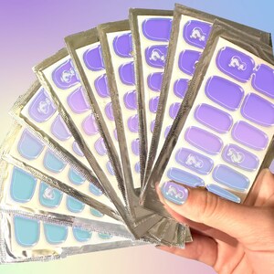 Könnte beinhalten: Ein Set mit acht Packungen Nagelfolien mit einem Farbverlauf in Blau- und Lilatönen. Jede Packung enthält mehrere Größen von Nagelfolien mit einem weißen Einhorn-Design.