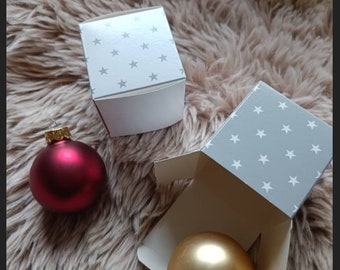Pode incluir: Duas caixas de presente brancas com um padrão de estrelas cinzentas, ao lado de uma bola de Natal vermelha e dourada. Os enfeites têm tampas douradas. Os itens estão em uma superfície fofa, marrom claro.