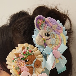 May include: Two embroidered horse hair clips, one brown and one pastel rainbow, with white and pink polka dot bows and white lace.
