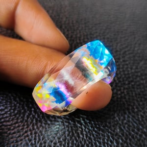 Puede incluir: Un anillo transparente iridiscente con un diseño facetado. El anillo tiene un brillo iridiscente azul, amarillo y rosa.