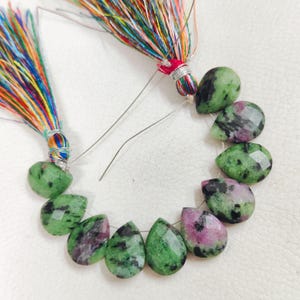 May include: A strand of faceted, pear-shaped ruby zoisite gemstones featuring a mottled pattern of vibrant green, black, and magenta-pink hues. The stones are strung on a thin wire and finished with two decorative, multi-colored rainbow tassels.