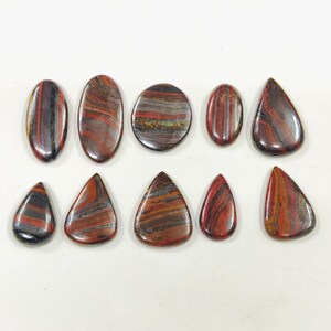 May include: A collection of ten polished gemstones in various shapes, including oval, teardrop, and triangular forms. The stones display a mix of red, brown, and black bands, creating a striped pattern. The gemstones are arranged on a white surface.