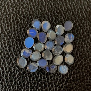 Peut inclure: Une collection de pierres précieuses ovales et irisées aux tons bleus et gris. Les pierres sont disposées sur une surface sombre et texturée, mettant en valeur leurs facettes polies et leurs variations naturelles. Les pierres varient en taille et en forme.