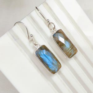 Puede incluir: Un par de pendientes colgantes con piedras preciosas de labradorita rectangulares de superficie facetada. Las piedras muestran destellos iridiscentes azules y dorados engastados en marcos de metal plateado con ganchos a juego.