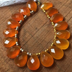 May include: A strand of orange carnelian gemstone beads, each bead is faceted and shaped like a teardrop. The beads are strung on a silver wire with gold spacers.
