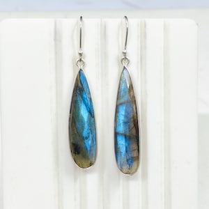 Puede incluir: Un par de pendientes en forma de lágrima con piedras de labradorita azul y verde iridiscentes. Las piedras están engastadas en un metal plateado y los pendientes tienen un diseño de gancho simple. Los pendientes se muestran sobre un fondo blanco.