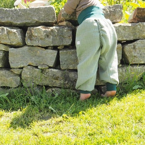 May include: A child wearing light green pants with a gathered back and a green waistband is standing near a stone wall. The child is looking down at the wall.