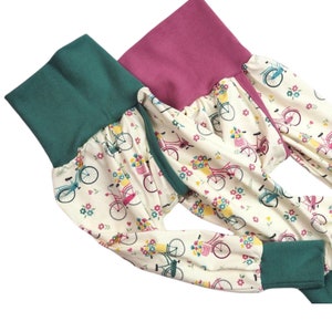 May include: Two pairs of cream-colored pants with a floral and bicycle print. One pair has a teal green waistband and cuffs, the other has a pink waistband and cuffs.