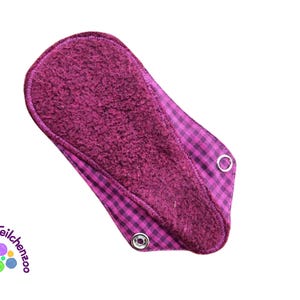 Puede incluir: Una compresa de tela reutilizable de color morado con un patrón de cuadros rosa y blanco en la parte posterior. La compresa tiene una textura suave y aterciopelada y está diseñada para la menstruación.