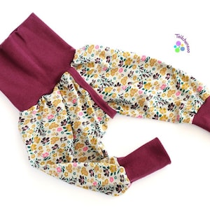 Puede incluir: Leggings de bebé en color burdeos y estampado floral con una cintura ancha. Las leggings tienen un estampado floral con flores rosas, amarillas y verdes sobre un fondo crema. La cintura es de color burdeos sólido.