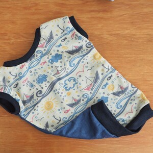May include: A light blue and beige baby onesie with a nautical theme. The onesie features a pattern of sailboats, fish, waves, and clouds. The onesie has a navy blue trim around the neck and armholes.