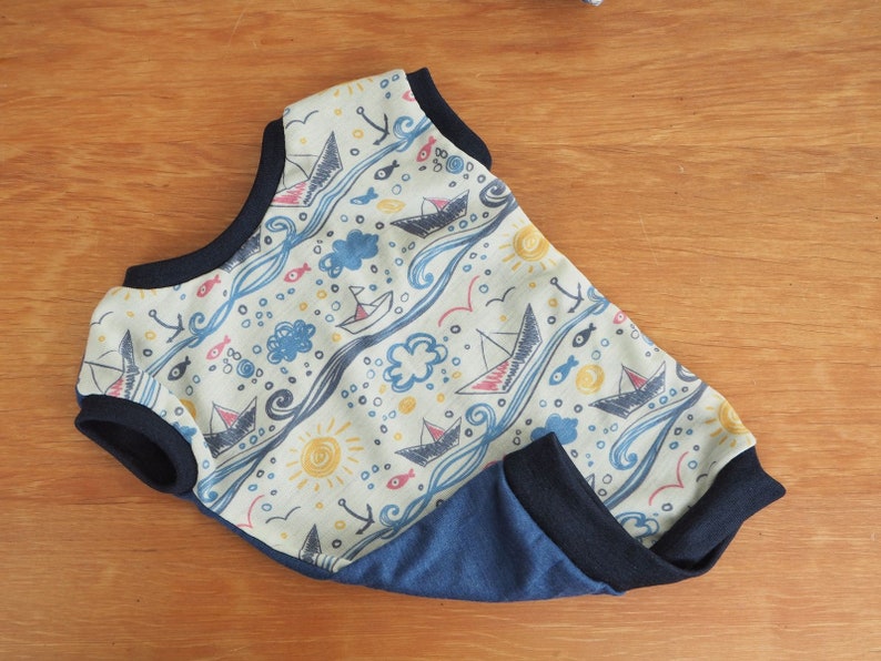 May include: A light blue and beige baby onesie with a nautical theme. The onesie features a pattern of sailboats, fish, waves, and clouds. The onesie has a navy blue trim around the neck and armholes.