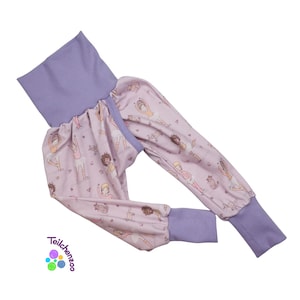 May include: Lilac and light pink baby pants with a ballet dancer pattern. The pants have a high, lilac waistband and matching cuffs. The fabric features illustrations of ballerinas in various poses. The brand name "Teilchenzoo" is visible.