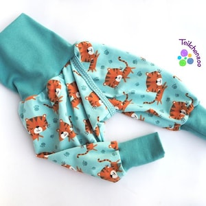 May include: Teal blue baby pants with a tiger print and paw prints. The pants have a fold-over cuff at the ankles and a fold-over cuff at the waist. The fabric is soft and comfortable. The pants are made by Teilchenzoo.