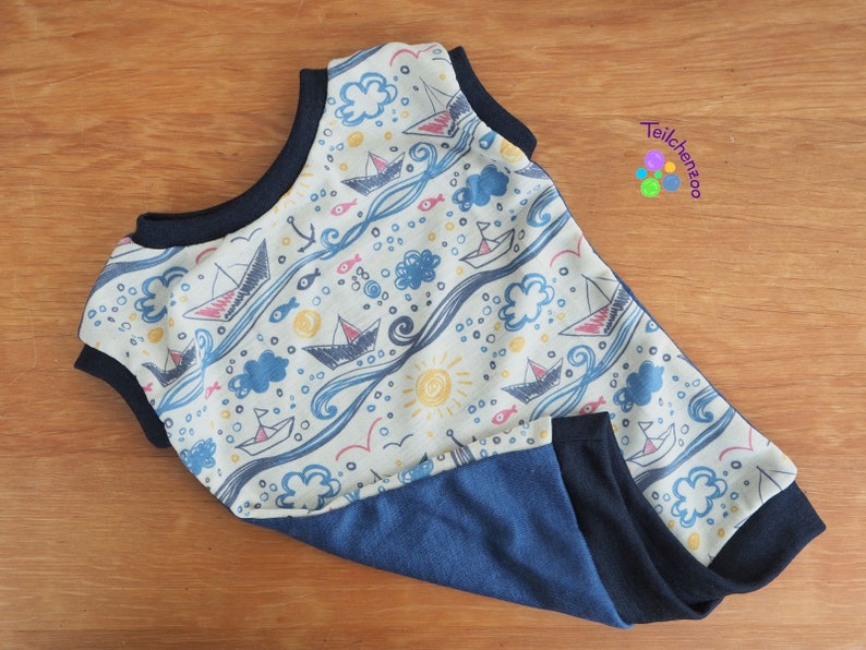 May include: A white sleeveless baby onesie with a blue and yellow nautical print featuring sailboats, waves, fish, and clouds. The onesie has a navy blue trim and a navy blue bottom.