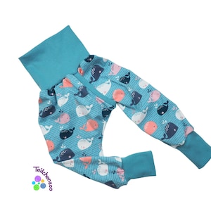 May include: Baby pants with a teal waistband and cuffs. The pants feature a light blue background with a pattern of whales in white, navy, and coral. The brand name "Teilchenzoo" is printed in the lower left corner.
