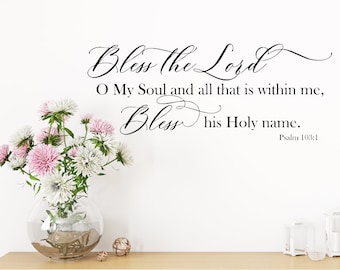 Pode incluir: Uma parede branca com uma citação de caligrafia preta que diz "Bless the Lord O My Soul and all that is within me, Bless His Holy name. Psalm 103:1". Um vaso com flores rosa e brancas está sobre uma mesa de madeira em primeiro plano.
