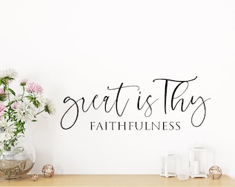 Pode incluir: Uma parede branca com uma citação de caligrafia preta que diz "great is thy faithfulness". A citação está centralizada na parede e é cercada por espaço em branco. Há flores em um vaso e objetos decorativos em uma prateleira de madeira abaixo da citação.