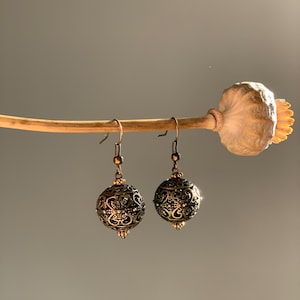May include: A pair of bronze-colored filigree ball earrings with intricate floral patterns. The earrings hang from delicate hooks, showcasing a vintage aesthetic. The earrings are suspended from a dried poppy pod.