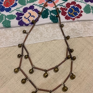May include: A double-strand necklace made of copper wire with small, round, brass bells hanging from it. The necklace is on a white fabric background with a floral pattern.