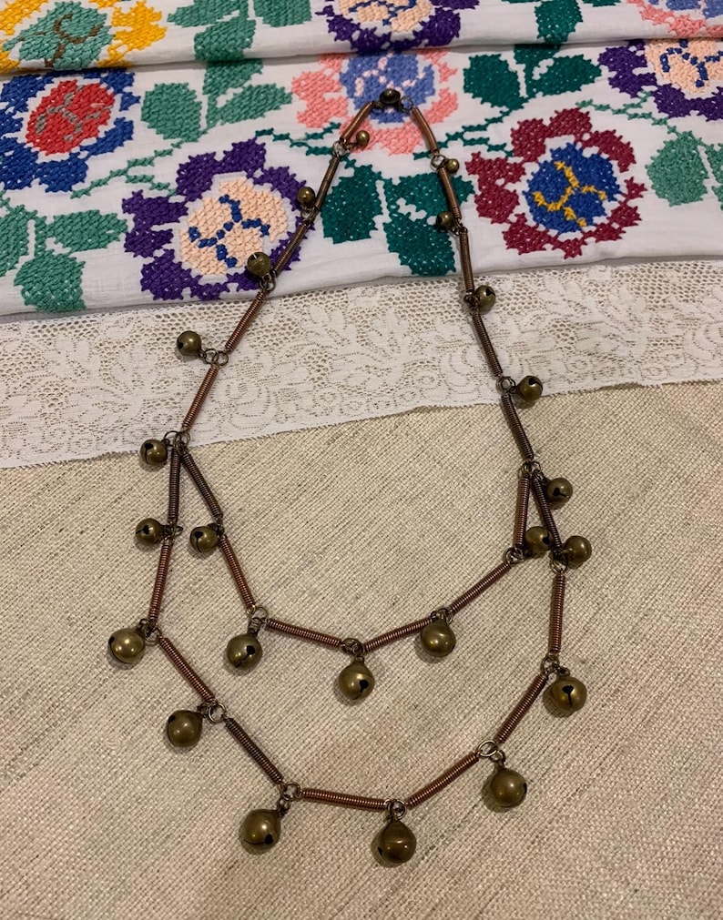 May include: A double-strand necklace made of copper wire with small, round, brass bells hanging from it. The necklace is on a white fabric background with a floral pattern.