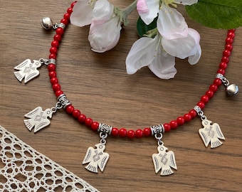 May include: A red beaded necklace with silver bird charms and bell accents. The necklace is displayed on a wooden surface with a white lace doily and a sprig of white apple blossoms.