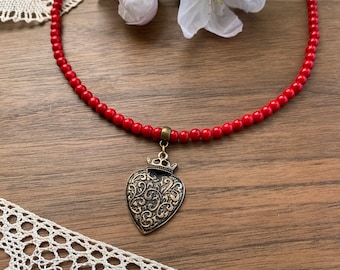 May include: A red beaded necklace with a heart-shaped pendant. The pendant is bronze-colored with an ornate design and a crown at the top. The necklace is displayed on a wooden surface with white flowers and lace.