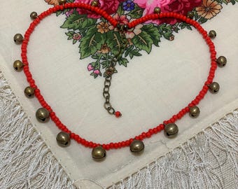 May include: A red beaded necklace with small gold bells hanging from it. The necklace is laid out in a heart shape on a white fabric with floral patterns.