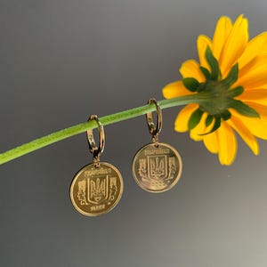 Puede incluir: Pendientes dorados con forma de moneda y diseño del escudo de armas ucraniano. Los pendientes redondos cuelgan de pequeños aros. El fondo es gris suave con una flor amarilla.