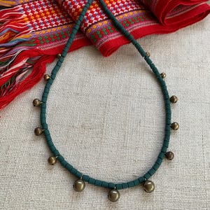 以下が含まれることがあります： 小さな金のベルがぶら下がった濃い緑色のビーズネックレス。ネックレスは薄い茶色の布地に置かれています。