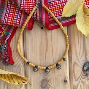 Puede incluir: Un collar artesanal con cuentas de madera marrón claro y tres campanas decorativas de metal. El collar se exhibe sobre una superficie de madera con un textil rojo estampado y hojas amarillas en el fondo.