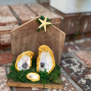 Può includere: Un presepe rustico in legno con conchiglie di ostrica dipinte d'oro. Le conchiglie sono disposte su un letto di muschio verde, con una piccola stella marina e muschio che accentuano il tetto di legno. La scena è ambientata su uno sfondo di mattoni.