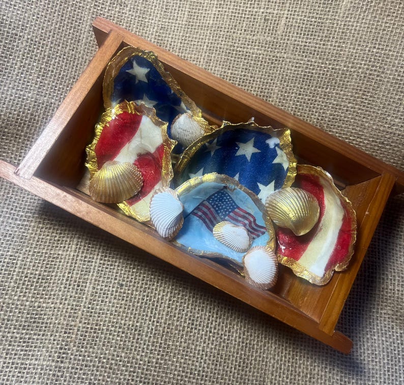 Puede incluir: Una bandeja de madera contiene varias conchas de ostra pintadas con motivos patri&oacute;ticos. Las conchas presentan rayas rojas y blancas, campos azules con estrellas blancas y banderas americanas. Incluye adornos dorados y conchas marinas.