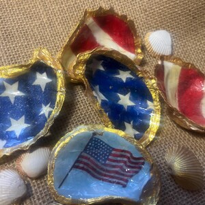 Puede incluir: Varias conchas de ostra decoradas con temas patri&oacute;ticos. Algunas conchas presentan un fondo azul con estrellas blancas, mientras que otras muestran rayas rojas y blancas. Los bordes est&aacute;n adornados con pintura dorada y peque&ntilde;as conchas marinas.
