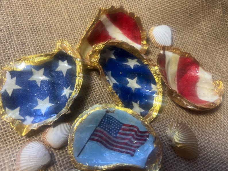 Puede incluir: Varias conchas de ostra decoradas con temas patri&oacute;ticos. Algunas conchas presentan un fondo azul con estrellas blancas, mientras que otras muestran rayas rojas y blancas. Los bordes est&aacute;n adornados con pintura dorada y peque&ntilde;as conchas marinas.