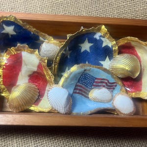 Puede incluir: Bandeja de madera llena de conchas de ostra pintadas. Las conchas est&aacute;n decoradas con el dise&ntilde;o de la bandera estadounidense, con rayas rojas y blancas, campos azules con estrellas blancas y una bandera estadounidense. Bordes dorados y conchas marinas.