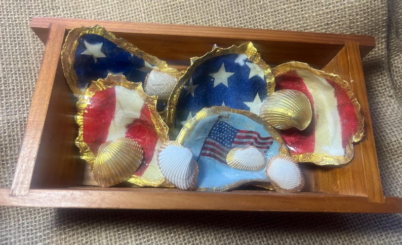Puede incluir: Bandeja de madera llena de conchas de ostra pintadas. Las conchas est&aacute;n decoradas con el dise&ntilde;o de la bandera estadounidense, con rayas rojas y blancas, campos azules con estrellas blancas y una bandera estadounidense. Bordes dorados y conchas marinas.
