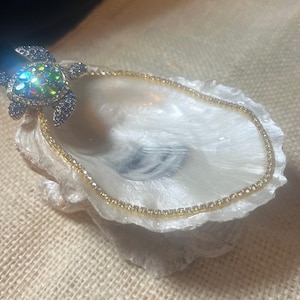 May include: A gold-toned chain bracelet with a sparkling, jeweled turtle charm. The turtle charm has a shimmering, iridescent center and dark gray accents. The bracelet rests in a white, textured oyster shell.