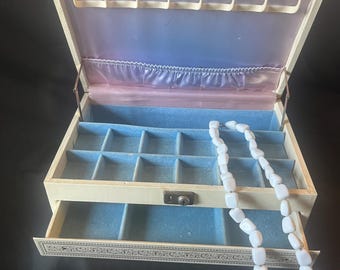 May include: A cream-colored jewelry box with a hinged lid, a pull-out drawer, and a light blue velvet interior. The lid features a row of hooks and a gathered pocket. A white beaded necklace rests inside the box, which has a metal latch.