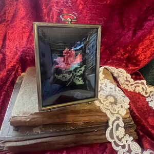 May include: A framed cross-stitch of a pink rose on black fabric. The frame is gold-colored with a small ring at the top. The frame rests on a stack of old books with a red velvet background and white lace trim.