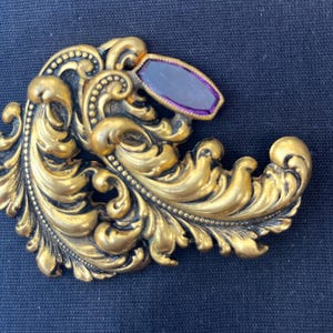 May include: Ornate gold-toned brooch with a swirling, leaf-like design. A rectangular, purple-colored stone is set at the top. The brooch has a vintage aesthetic, with detailed scrollwork and a polished finish.