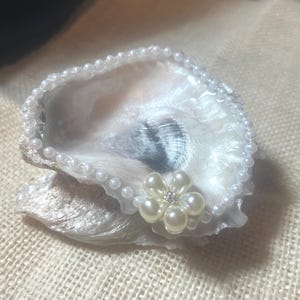 May include: A decorative oyster shell, painted white with a pearlescent sheen, is adorned with a delicate border of small pearls. A pearl flower with a rhinestone center accents the shell's edge, creating an elegant coastal theme.