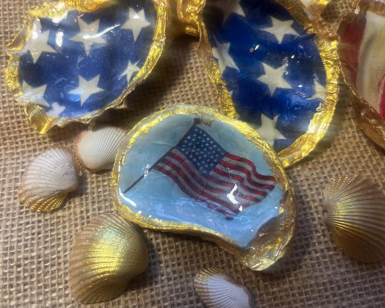 Puede incluir: Varias conchas de ostra decoradas con motivos patri&oacute;ticos. Algunas conchas presentan un fondo azul con estrellas blancas, mientras que otras muestran un dise&ntilde;o de la bandera estadounidense. Las conchas tienen bordes dorados y est&aacute;n rodeadas de conchas doradas y naturales.