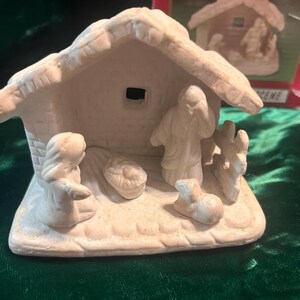 May include: A small, unpainted ceramic nativity scene figurine featuring a stable structure with a thatched roof, a central manger, and figures of three individuals and a small animal. The piece is off-white with a matte, textured finish.
