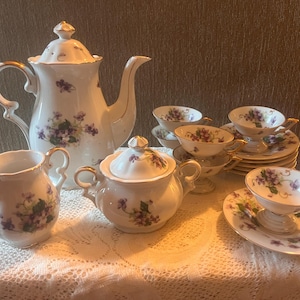 May include: A white porcelain tea set featuring a delicate violet floral pattern and gold trim. The set includes a tall teapot with a lid, a creamer, a lidded sugar bowl with two handles, and several teacups with matching saucers.