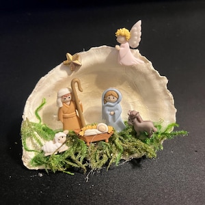 May include: A miniature Christmas nativity scene, featuring figurines of Mary, Joseph, baby Jesus, an angel, a lamb, and a donkey, set within a seashell. The figures are in muted tones, with a star above. Green moss adds a natural touch.